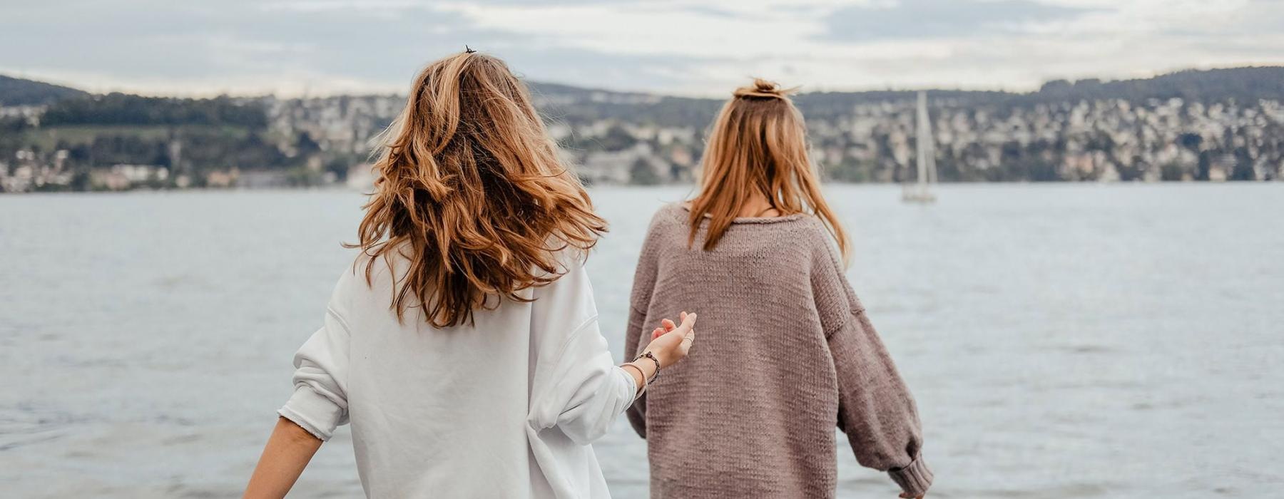two women walk to the edge of a dock, in a body of water, looking toward the city on the other shore